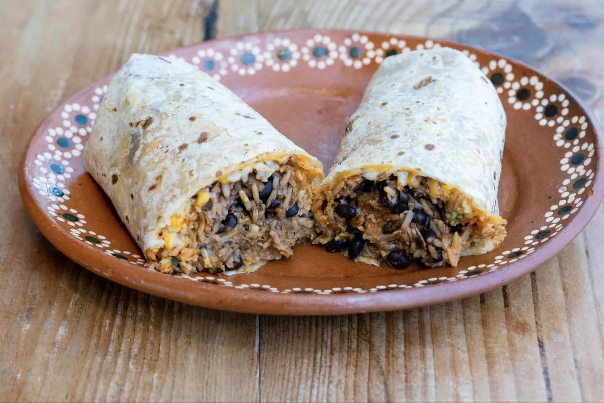 A burrito cut in half, filled with rice, black beans, shredded meat, and cheese, on a clay plate