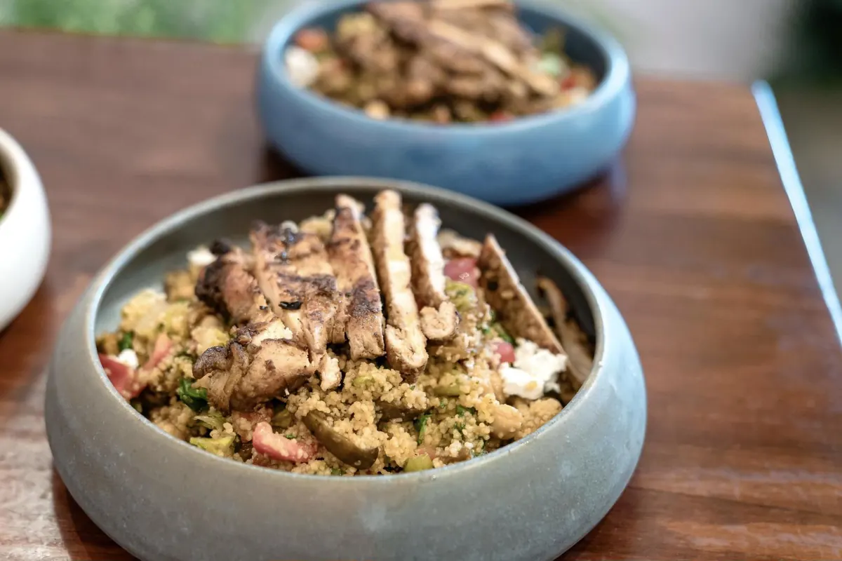 A bowl of quinoa salad topped with sliced grilled chicken, tomato, and white cheese, with more bowls behind