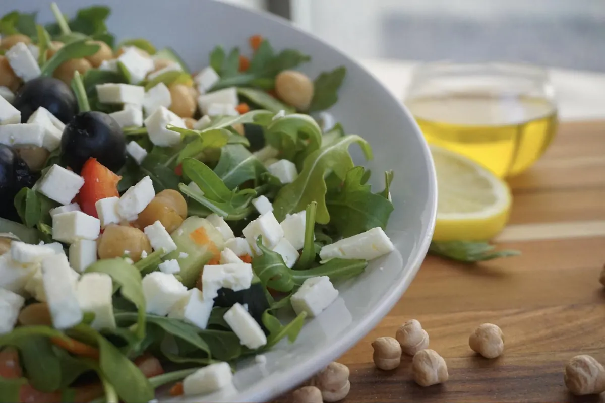 A bowl of salad with chickpeas, feta cubes, arugula, black olives, and red pepper, with olive oil and lemon behind
