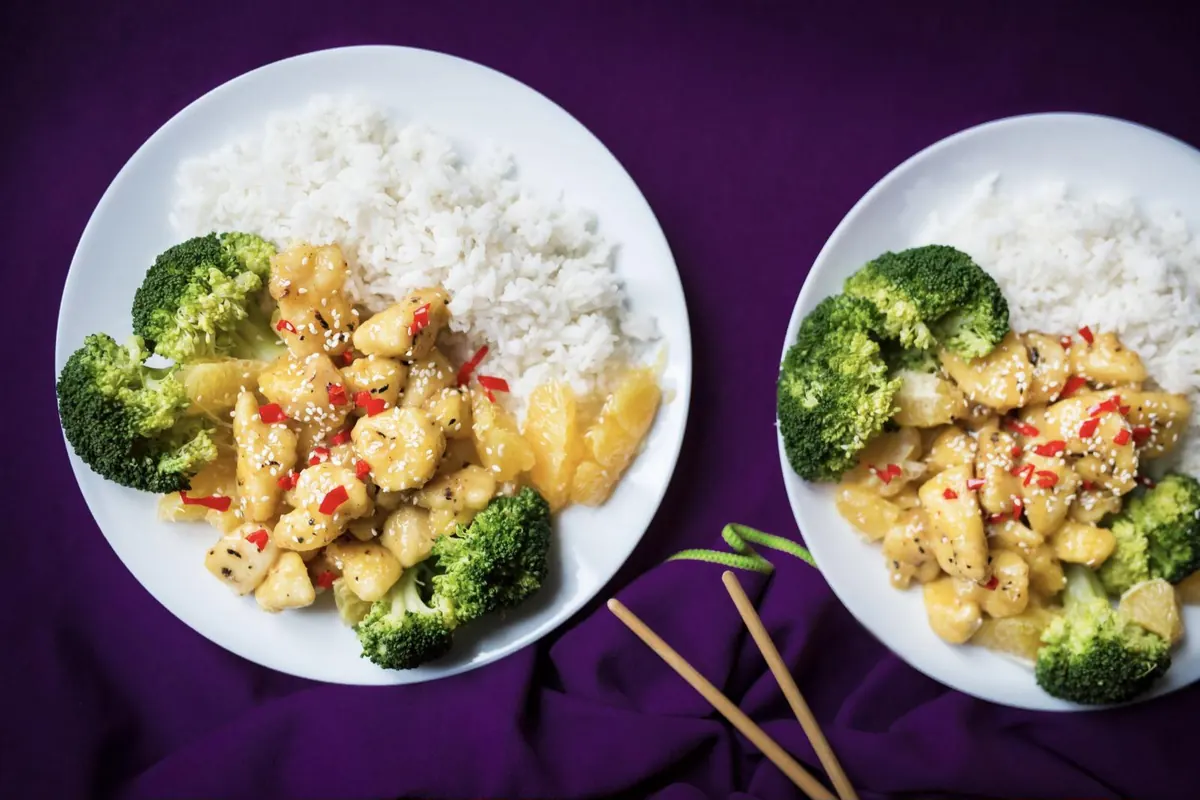 Two plates of glazed chicken pieces with sesame seeds, broccoli, orange segments, and white rice
