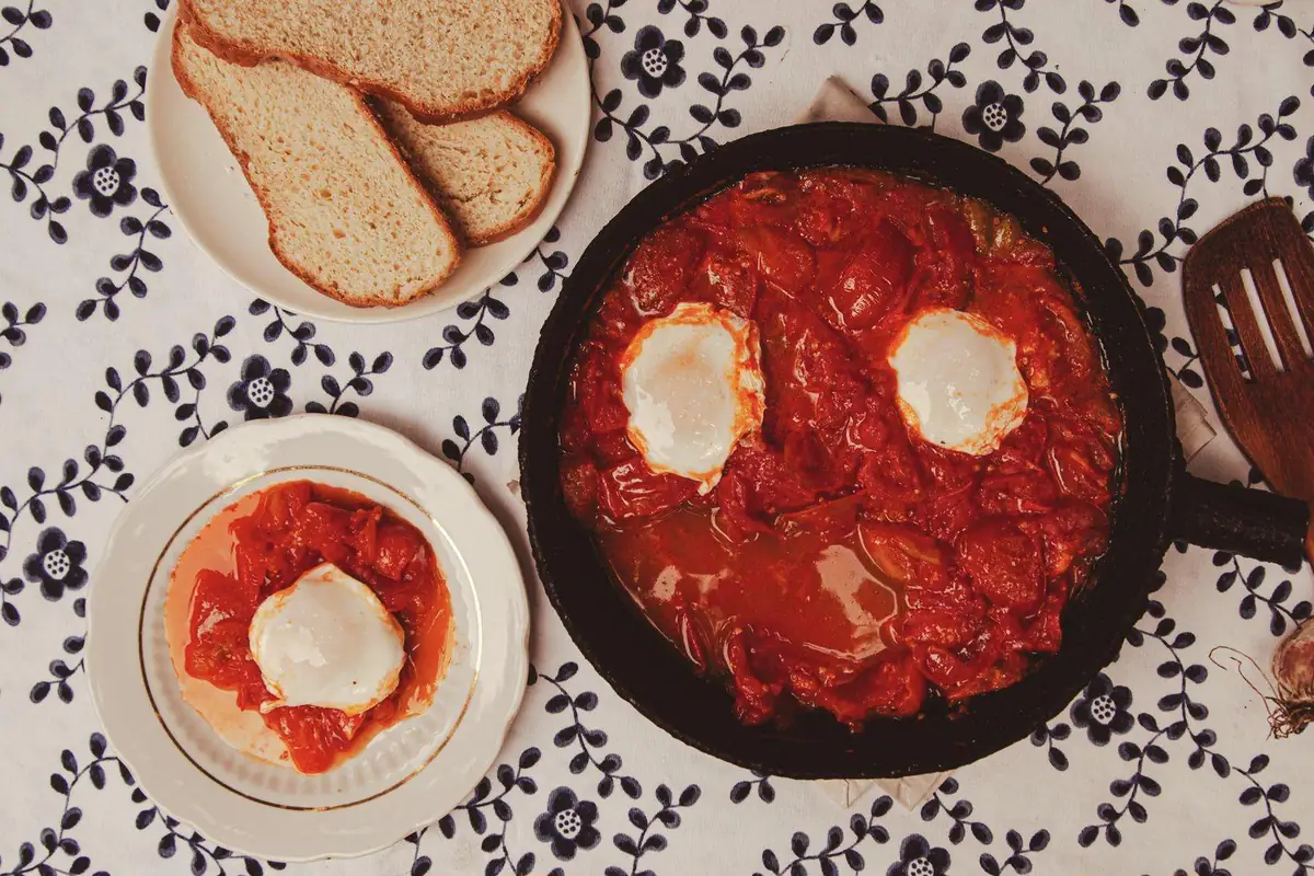 Shakshuka in a cast-iron skillet with two eggs, a served plate with one egg, and sliced bread on a floral tablecloth