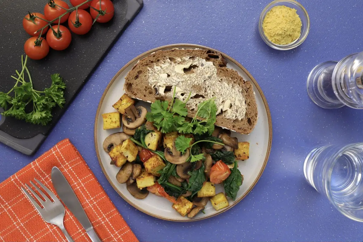 A plate of golden tofu cubes with mushrooms, spinach, and tomato beside a slice of buttered bread, with cherry tomatoes and parsley nearby